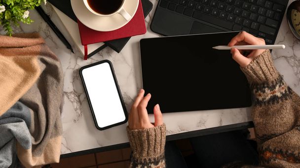 Vue du dessus des mains de créateurs féminines travaillant avec une tablette numérique sur un espace de travail moderne avec une pile de vêtements, un écran vide pour smartphone et des fournitures - Photo, image