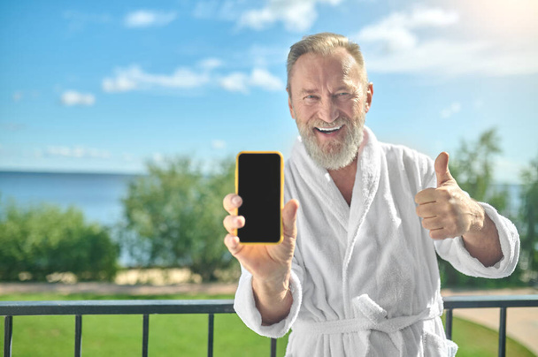 Male in the bathrobe showing his smartphone on the balcony - Φωτογραφία, εικόνα