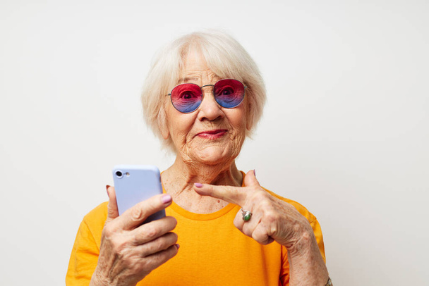 smiling elderly woman in fashionable glasses with a smartphone in hand light background - Фото, изображение