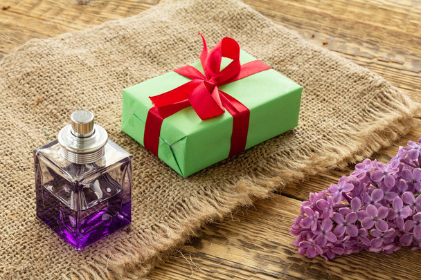 Egy üveg parfüm, egy zöld ajándékdoboz zsákvásznon és orgonavirág a fa deszkákon. Fogalom az ajándékozásról az ünnepekkor. Felülnézet. - Fotó, kép
