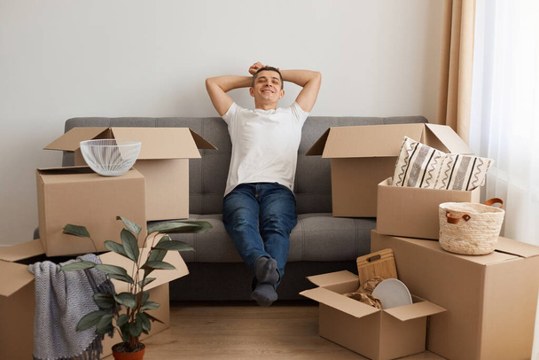 Happy satisfied relaxed man wearing white T-shirt and jeans sitting on sofa surrounded with pile of packages, looking at camera with smile, keeping hands behind head, being glad to move to a new flat. - Photo, Image