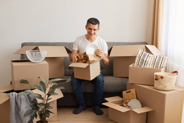 Indoor shot of optimistic Caucasian man wearing white T-shirt and jeans sitting on cough surrounded with pile of packages, unpack personal stuff from carton boxes, expressing happiness. - Foto, Imagem
