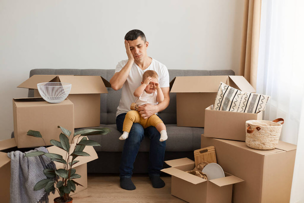 Portrait of tired exhausted man wearing white t-shirt and jeans sitting on sofa with his baby daughter, moving to a new home, posing with crying child, suffering terirble headache, keeps eyes closed - Фото, изображение