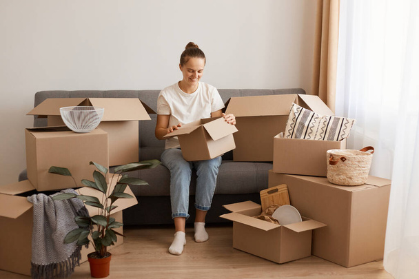 Indoor shot of positive smiling woman wearing white t-shirt and jeans sitting on sofa in a new house or flat after relocating, holding cardboard box, looking inside, unpacking packages. - Photo, image