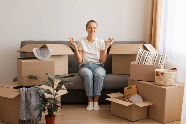 Portrait of smiling satisfied woman wearing white t-shirt and jeans sitting on cough in a new apartment after moving, looking at camera, posing in mudra position, surrounded with belongings in boxes. - Foto, immagini