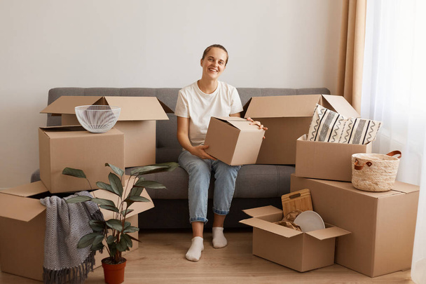 Portrait of attractive happy woman wearing white t-shirt and jeans sitting on cough in a new apartment after moving, looking at camera with smile and happy expression, holding cardboard box. - Photo, Image