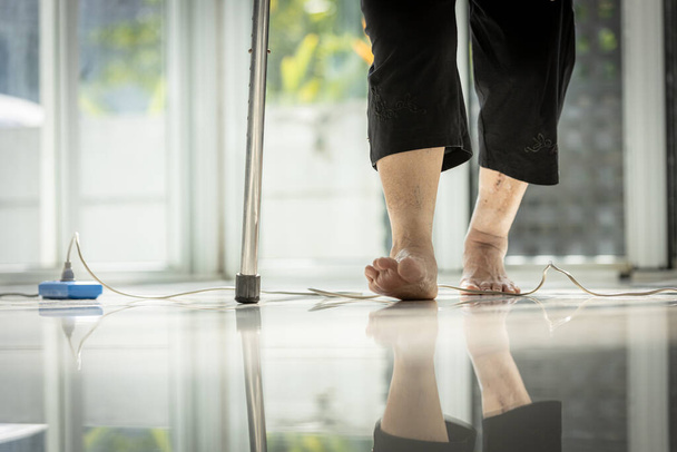 Close-up, benen van bejaarden met stok ter ondersteuning van haar wandeling, voeten stappen over een stopcontact, risico van struikelen over netsnoer op de vloer, concept van gevaar en ongeval in huis - Foto, afbeelding