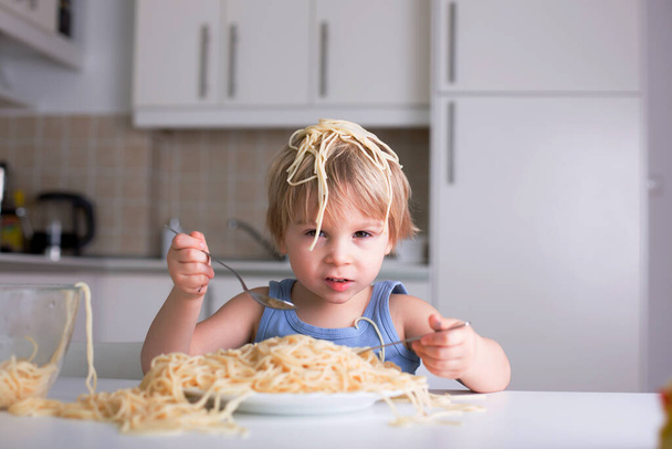 Little blond boy, toddler child, eating spaghetti for lunch and making a mess at home in kitchen - Foto, Imagem
