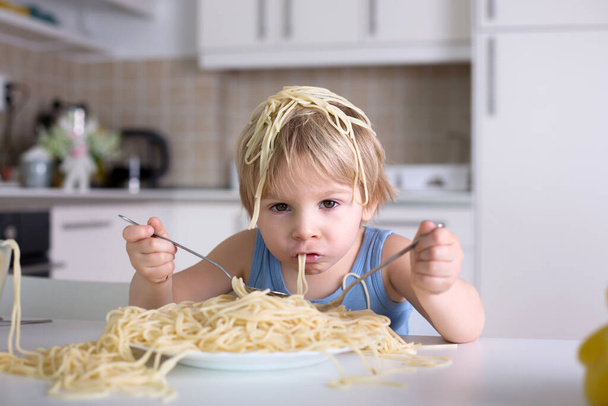 Little blond boy, toddler child, eating spaghetti for lunch and making a mess at home in kitchen - Photo, image