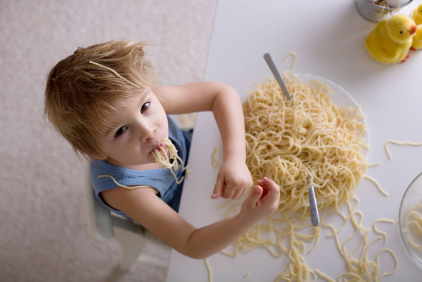 Little blond boy, toddler child, eating spaghetti for lunch and making a mess at home in kitchen - Fotoğraf, Görsel
