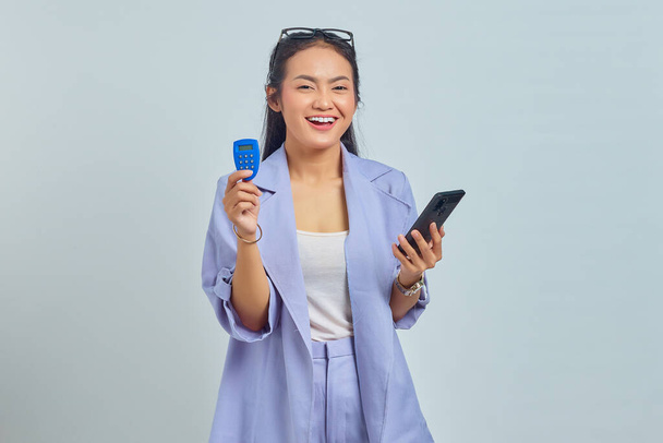Portret wesołej młodej Azjatki trzymającej żeton bankowy i trzymającej smartfona na białym tle. koncepcja przelewu środków przez Internet - Zdjęcie, obraz