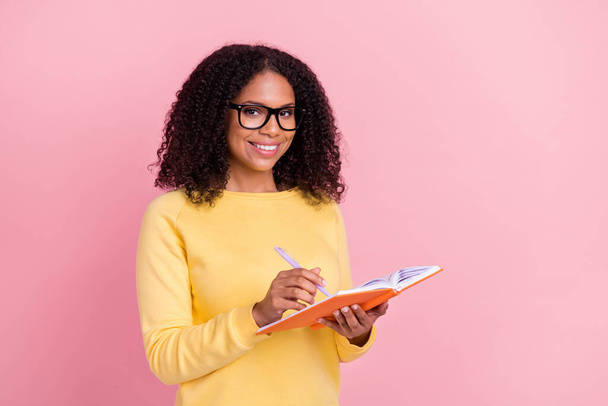 Photo of adorable shiny dark skin woman wear yellow sweater spectacles writing copybook isolated pink color background - 写真・画像
