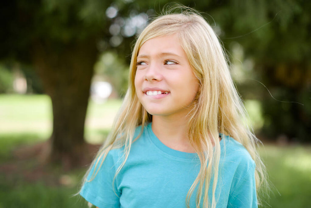 Increíble niña caucásica usando camiseta azul de pie al aire libre mordiendo el labio y buscando difícil de espacio vacío. - Foto, imagen