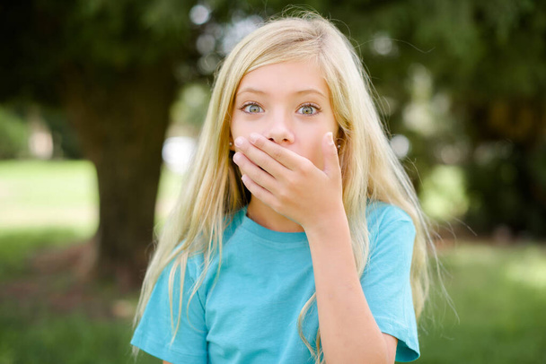 Emocional niña caucásica usando camiseta azul de pie al aire libre jadeos de asombro, cubre la boca abierta con la palma, se ve sorprendido por la cámara. Concepto de reacción - Foto, Imagen