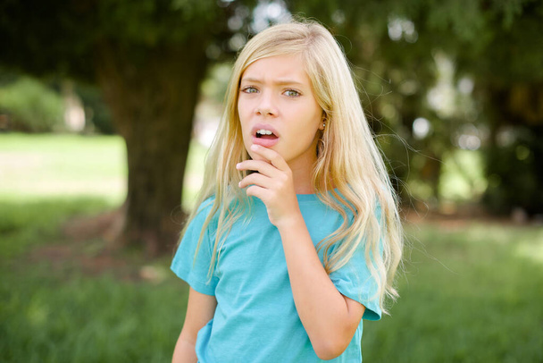 Nerwowy, zdziwiony, biały dzieciak, dziewczynka w niebieskiej koszulce, stojąca na zewnątrz, otwiera usta z zaskoczenia, reaguje na nagłe wiadomości.. - Zdjęcie, obraz
