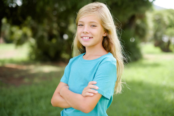 Biała dziewczynka w niebieskiej koszulce stojąca na zewnątrz uśmiechnięta z skrzyżowanymi rękami patrząca w kamerę. Osoba pozytywna. - Zdjęcie, obraz