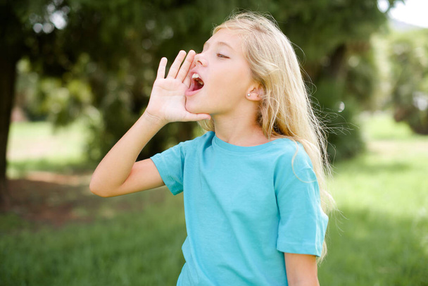 Маленькая белая девочка в голубой футболке, стоящая на улице и громко кричащая, прижавшись к губам. Концепция коммуникации. - Фото, изображение