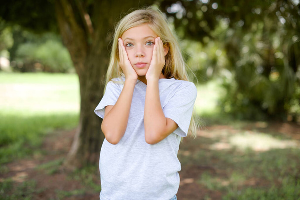 Kaukasisches kleines Mädchen mit weißem T-Shirt, das draußen steht Müde Hände, die Gesicht, Depression und Traurigkeit verdecken, verärgert und irritiert wegen des Problems - Foto, Bild