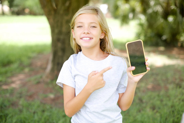Χαμογελώντας Καυκάσιος μικρό κορίτσι φορώντας λευκό T-shirt στέκεται σε εξωτερικούς χώρους Mock up αντίγραφο χώρο. Δάχτυλο δείκτη δείχνοντας στο κινητό τηλέφωνο με κενή κενή οθόνη - Φωτογραφία, εικόνα