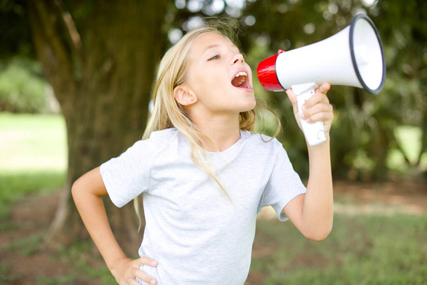Funny beautiful Caucasian little kid girl wearing whiteT-shirt standing outdoors People sincere emotions lifestyle concept. Mock up copy space. Screaming in megaphone. - Foto, Imagem