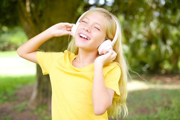 όμορφο λευκό κοριτσάκι φορώντας κίτρινο T-shirt στέκεται σε εξωτερικούς χώρους χαμόγελα αισθάνεται γενικά πολύ χαρούμενος ακούει αγαπημένο μουσικό κομμάτι μέσω ασύρματων ακουστικών κλείνει τα μάτια. - Φωτογραφία, εικόνα