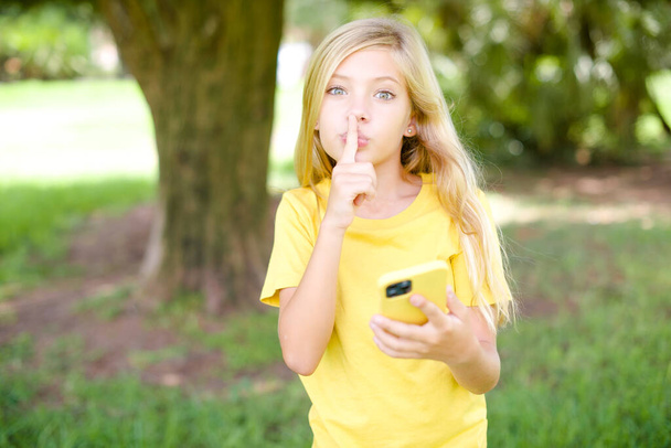 schöne kaukasische kleine Mädchen tragen gelbes T-Shirt im Freien stehend mit modernen Gadget fragen, nicht Geheimnisse zu erzählen - Foto, Bild
