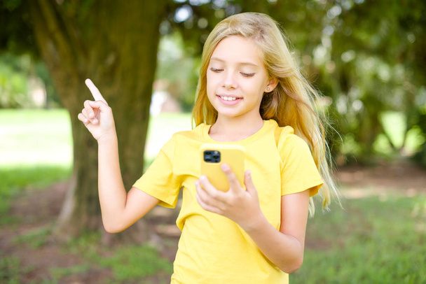 Χαμογελώντας όμορφο λευκό κοριτσάκι φορώντας κίτρινο T-shirt στέκεται σε εξωτερικούς χώρους δείχνοντας το δάχτυλο σε κενό χώρο κρατώντας το τηλέφωνο στο ένα χέρι - Φωτογραφία, εικόνα