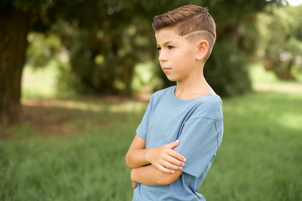 hermoso caucásico pequeño niño vistiendo azul camiseta de pie al aire libre cruzar las manos mirar espacio de copia - Foto, Imagen