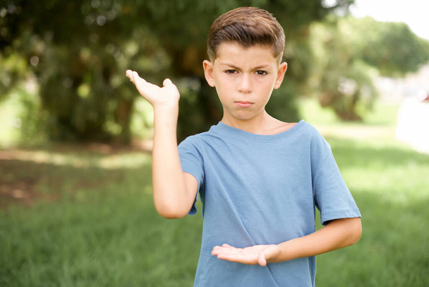 Güzel, beyaz, mavi tişörtlü bir çocuk iki eliyle dışarıda dikilip tuhaf bir şey gösteriyor ve "Bu ne bilmiyorum". diyor. Reklam kavramı. - Fotoğraf, Görsel
