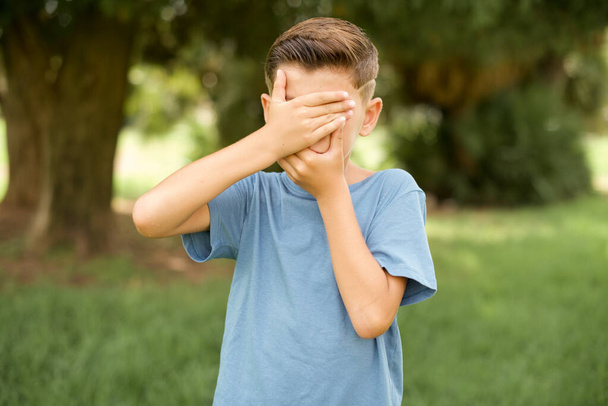 beautiful Caucasian little kid boy wearing blue T-shirt standing outdoors  Covering eyes and mouth with hands, surprised and shocked. Hiding emotions. - Photo, Image
