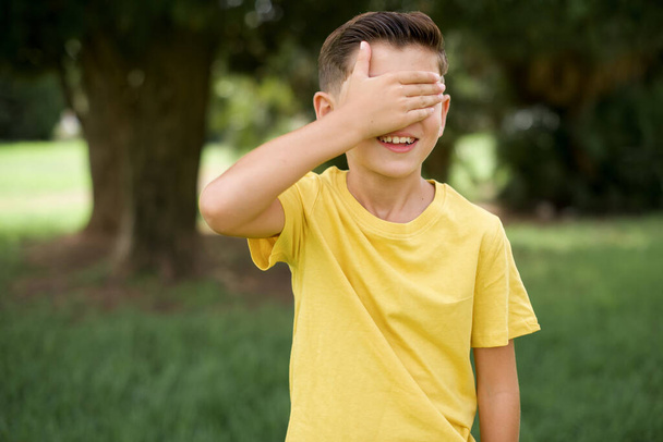 wunderschöner kaukasischer kleiner Junge in gelbem T-Shirt, der im Freien steht, lächelt und zur Überraschung mit der Hand auf dem Gesicht die Augen verdeckt. Blindes Konzept. - Foto, Bild