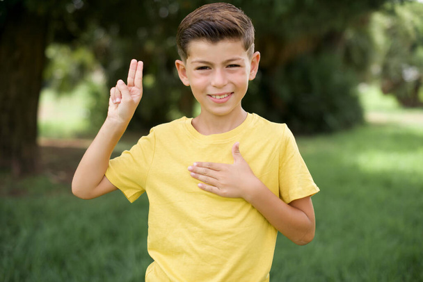 mooi blank jongetje jongen dragen geel T-shirt buiten staan glimlachen vloeken met de hand op de borst en vingers omhoog, het maken van een trouw belofte eed. - Foto, afbeelding