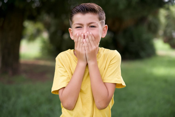 Vivacious lindo caucasiano menino vestindo camiseta amarela em pé ao ar livre, risos alegremente, cobre a boca, tem riso natural, ouve história positiva ou anedota engraçada - Foto, Imagem