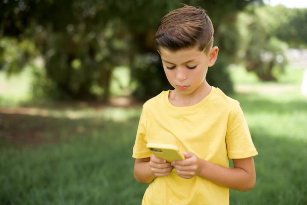 Excited beautiful Caucasian little kid boy wearing yellow T-shirt standing outdoors winking and eye hold smart phone use read social network news - Photo, Image