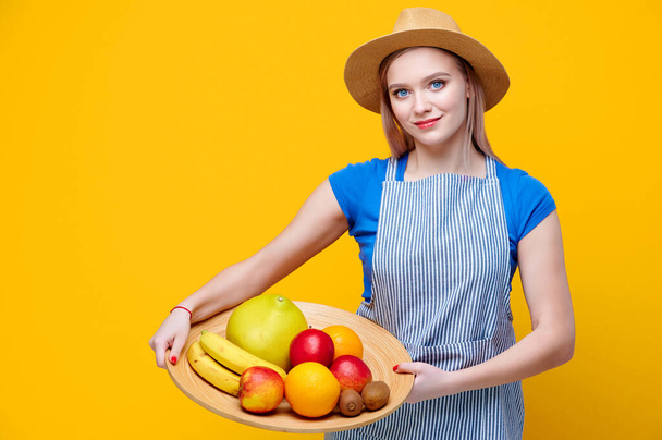 若い女性庭師が果物バナナリンゴオレンジキウイグレープフルーツを持っている木の板に藁帽子と黄色の背景にエプロンを身に着けています - 写真・画像