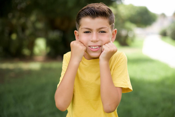 Happy beautiful Caucasian little kid boy wearing yellow T-shirt standing outdoor keeps fists on cheeks smile wide and has positive expression being in good mood - Foto, afbeelding