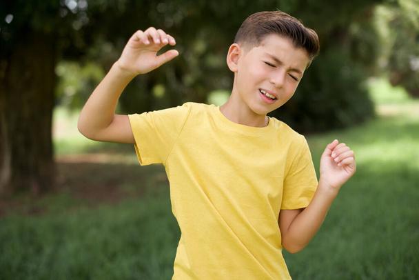 Photo de joyeux petit garçon caucasien vêtu de T-shirt jaune debout à l'extérieur a du plaisir et danse vêtements insouciants être d'humeur parfaite fait des mouvements. Passe du temps libre sur disco partie - Photo, image