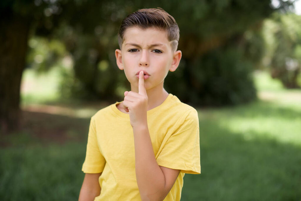 Sorprendido hermoso niño caucásico niño usando camiseta amarilla de pie al aire libre hace gesto de silencio, mantiene el dedo sobre los labios y se ve misterioso en la cámara - Foto, imagen
