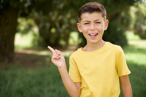 beautiful Caucasian little kid boy wearing yellow T-shirt standing outdoors points aside on copy blank space. People promotion and advertising concept - Fotoğraf, Görsel