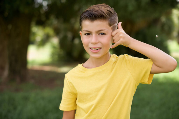 schöner kaukasischer kleiner Junge mit gelbem T-Shirt, der draußen steht, macht Telefongeste, sagt, ruf mich wieder an, hat einen fröhlichen Ausdruck. - Foto, Bild