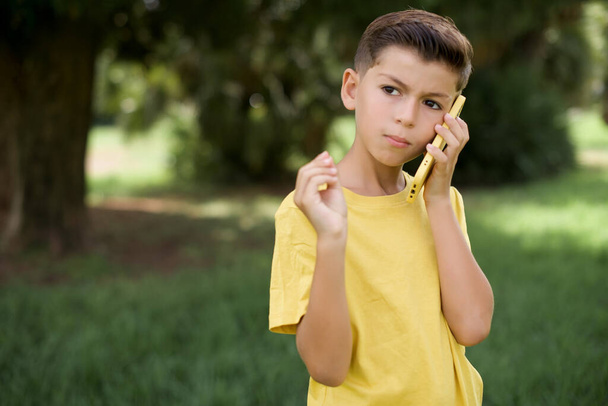 beautiful Caucasian little kid boy wearing yellow T-shirt standing outdoors speaks on mobile phone spends free time indoors calls to friend. - Photo, Image