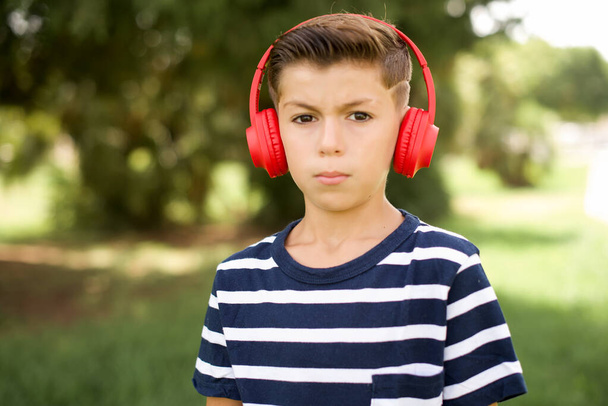 Poważny niezadowolony piękny biały chłopiec w paski T-shirt stojący na zewnątrz wygląda na zaskoczonego, gdy kamera jest zły nosi słuchawki stereo słucha muzyki podczas spaceru po ulicy - Zdjęcie, obraz