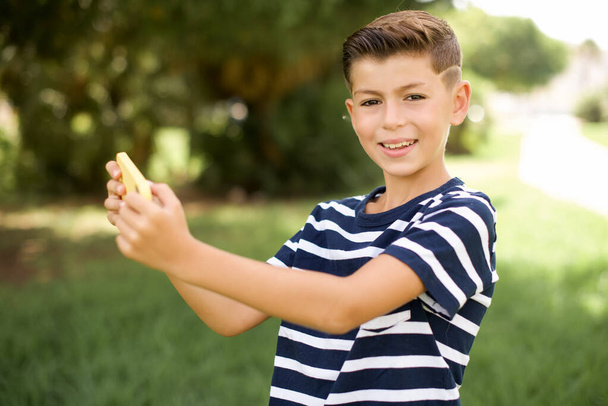 Niza adicto alegre hermoso niño caucásico pequeño usando camiseta a rayas de pie al aire libre usando gadget red de juego - Foto, imagen