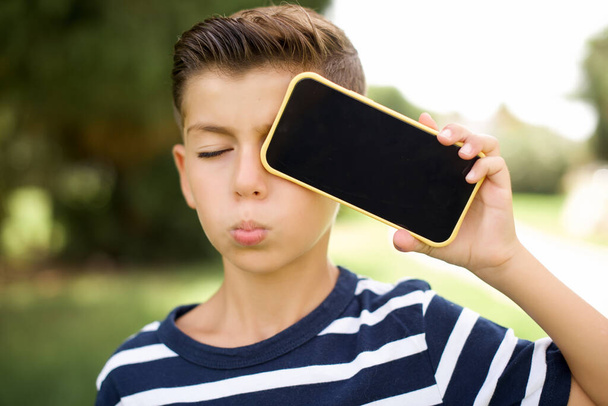 Adorable beautiful Caucasian little kid boy wearing striped T-shirt standing outdoors holding modern device covering eye with lips pouted - Photo, Image