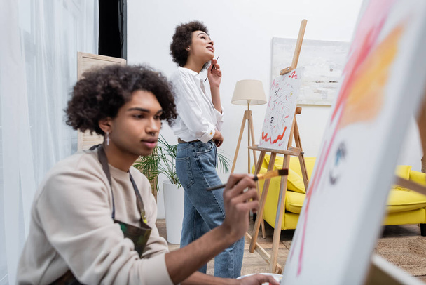 Мрійлива афро-американська жінка, що тримає пензель біля полотна і розмитий хлопець малює вдома.  - Фото, зображення