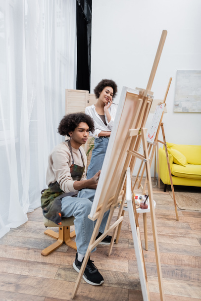 Mujer afro americana positiva mirando novio en delantal pintura sobre lienzo en casa  - Foto, Imagen