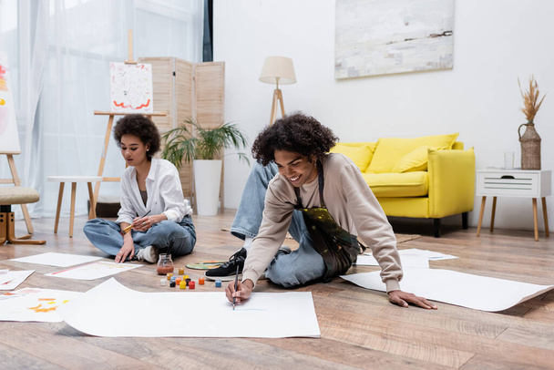 Positive african american man painting on paper on floor near girlfriend at home  - Photo, Image
