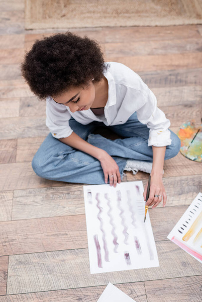 Overhead view of african american woman painting while sitting on floor at home  - Foto, Imagem