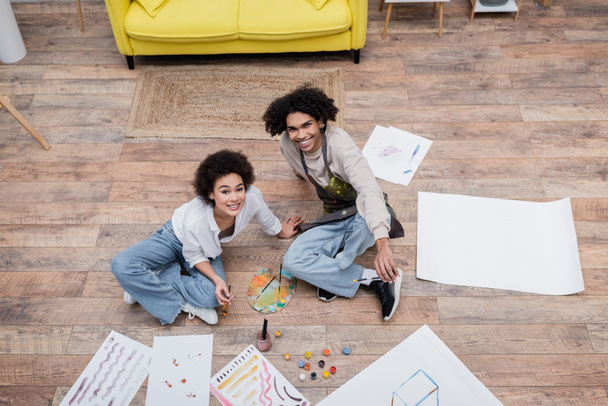 Top view of cheerful african american couple looking at camera near papers and paints on floor  - Photo, Image