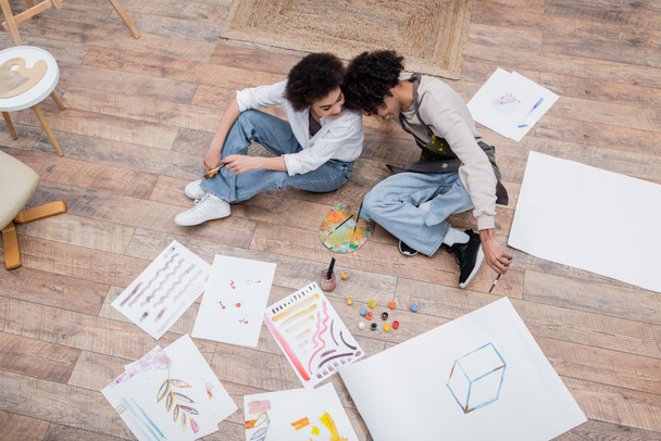 Overhead view of cheerful african american couple holding paintbrushes near papers and paints on floor  - Foto, Imagem